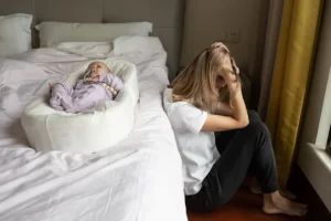 Kvinna sitter vid sidan av sängen ledsen. Ett barn ligger på sängen