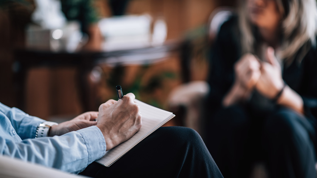 Psykiatriker skriver anteckningar i ett block med en patienten mittemot sig.