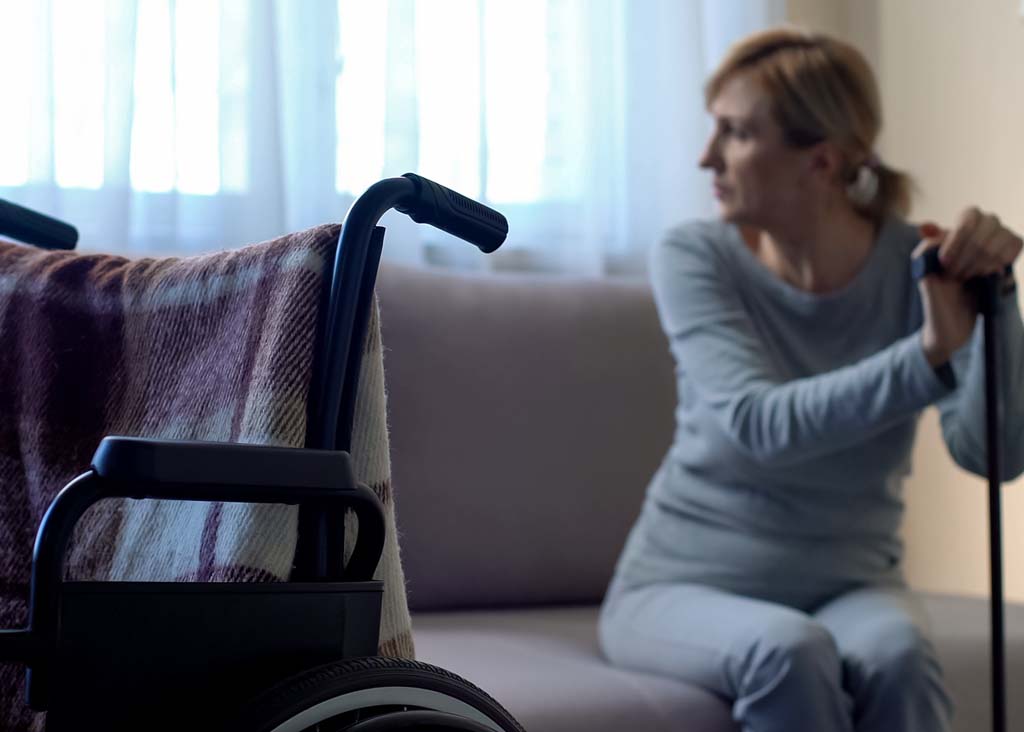 Kvinna sitter i en soffa och stödjer sig på en käpp en rullstol syns i förgrunden