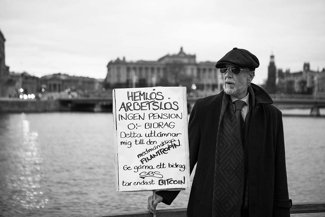 Finklädd man står med ett plakat bakom honom syns riksdagshuset. På plakatet står: Hemlös, arbetslös, ingen pension, noll kronor i bidrag ge gärna ett bidrag.