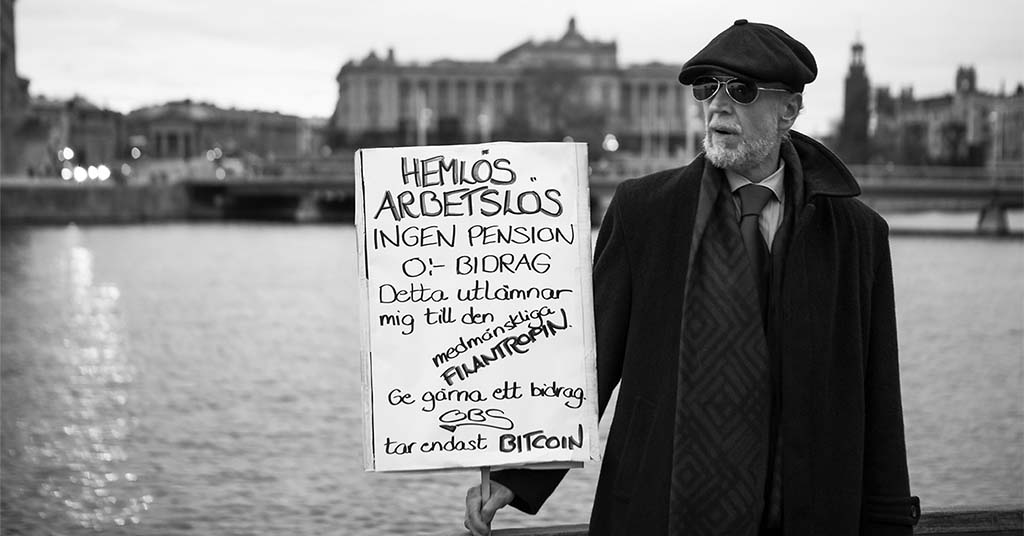 Finklädd man står med ett plakat bakom honom syns riksdagshuset. På plakatet står: Hemlös, arbetslös, ingen pension, noll kronor i bidrag ge gärna ett bidrag.